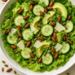 Close-up of a fresh green salad with soft lettuce, cucumber slices, avocado, Parmesan, microgreens, roasted tamari almonds, and pepitas in a white bowl, styled with scattered seeds and nuts for decoration.